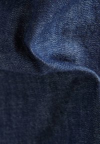 Primer plano de una tela de mezclilla azul oscura que muestra la textura y el patrón de tejido detallado con pliegues y sombras visibles.