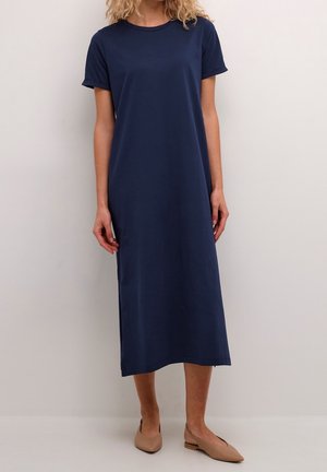 Frau trägt ein marineblaues Midi-Kleid mit kurzen Ärmeln und beigefarbene spitze flache Schuhe, steht vor einem schlichten hellen Hintergrund.