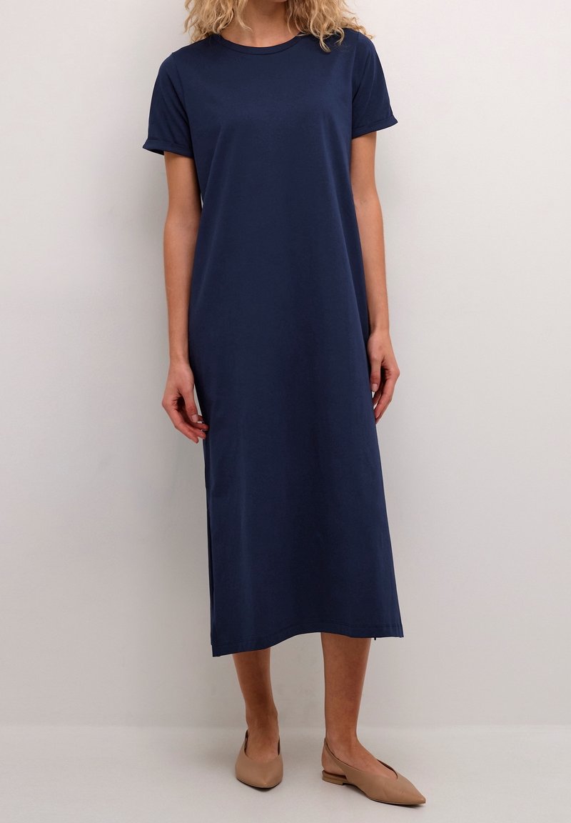 Femme portant une robe midi bleu marine à manches courtes et des ballerines pointues beige, debout devant un fond clair uni.