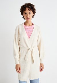 Beige stickad cardigan med ett strukturmönster, öppen front och knytning i midjan. Långa ärmar och en avslappnad passform, tillsammans med en rosa stickad topp.