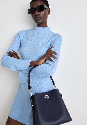 Vrouw met zwarte rechthoekige zonnebril en een lichtblauw geribbelde sweaterjurk, met een donkerblauwe leren handtas in haar hand.