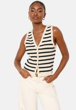 Woman wearing a sleeveless black and white striped knit vest with gold buttons and cream high-waisted pants, hands in pockets.
