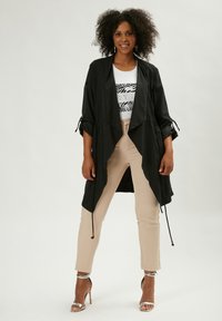 Cárdigan negro, ligero y drapeado con mangas enrolladas y lazos ajustables. Llevado sobre una camiseta blanca y pantalones beige ajustados. Sandalias de tacón plateadas.