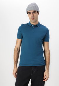Tyrkisk grønn polo med korte ermer, krage og tre knapper. Brukt sammen med mørke jeans og en lys grå strikket lue.