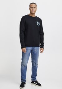 Felpa nera con collo a giro e logo blu sul petto, abbinata a jeans blu sbiaditi e scarpe da ginnastica nere con strisce bianche.