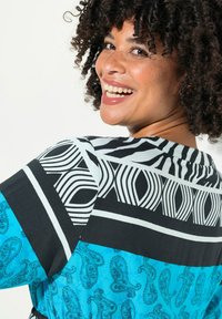 Pestrobarevná blůza s černobílým geometrickým vzorem a modrým paisley vzorem. Má volný střih a krátké rukávy.