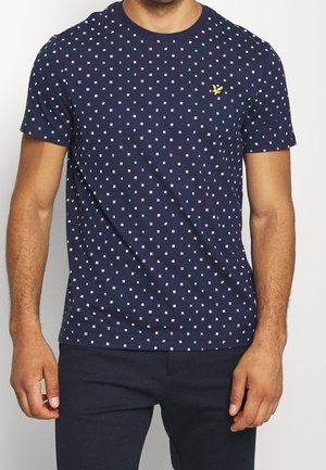 Mann trägt ein marineblaues T-Shirt mit einem kleinen weißen und türkisfarbenen Quadratmuster, kombiniert mit dunkler Hose, zugeschnitten am Hals und auf halber Oberschenkelhöhe.