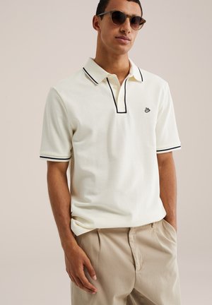 Uomo che indossa una polo color crema con bordino nero e pantaloni beige, in piedi con una mano in tasca e occhiali da sole su sfondo neutro.