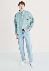 Cămașă cu dungi verzi și albe, cu un buzunar frontal, asortată cu blugi din denim de un albastru deschis și adidași albi.