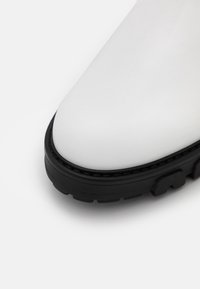 Gros plan sur la pointe d'une chaussure en cuir blanc avec une semelle en caoutchouc noire épaisse et un motif de crampons texturé.