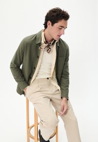 Olivenfarbene Button-Down-Jacke über einem cremefarbenen gerippten Shirt, kombiniert mit beigen Hosen. Ein gemusterter Schal unterstreicht den Look.