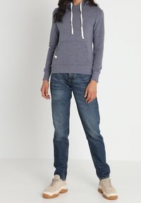 Szara bluza z kapturem z kremowymi sznurkami, kieszenią kangurką, w połączeniu z niebieskimi dżinsami i beżowymi trampkami. Gładka faktura, swobodny krój, prosty design.