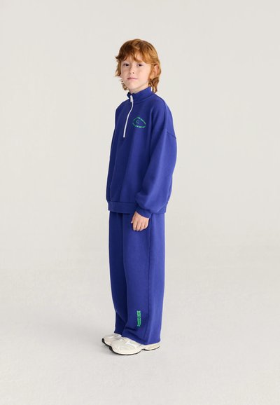 Enfant aux cheveux roux debout portant un survêtement bleu avec des baskets blanches sur un fond clair uni.