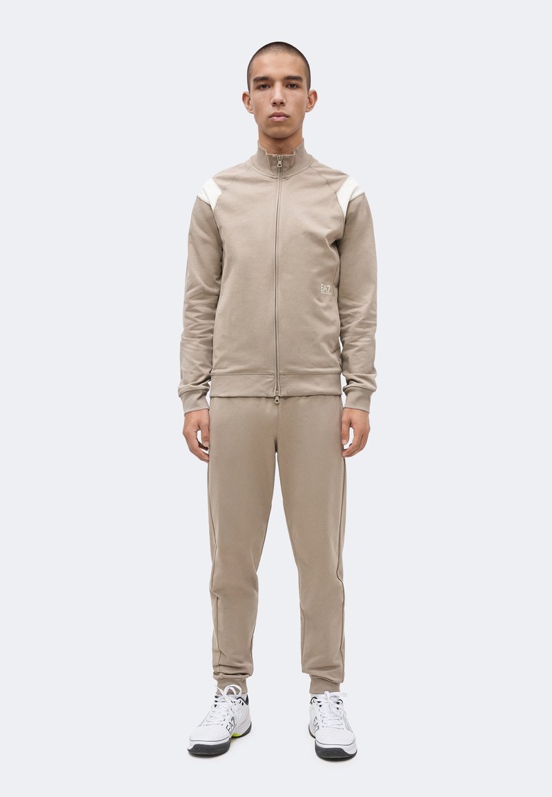 Άνδρας που φοράει μπεζ φόρμα με φερμουάρ, λευκές πλάκες στους ώμους και λευκά αθλητικά παπούτσια, μπροστά σε απλό ανοιχτόχρωμο φόντο.