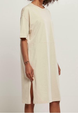 Vrouw draagt een loszittende beige jurk tot op de knie met korte mouwen en een zijsplit, staand tegen een effen lichte achtergrond.