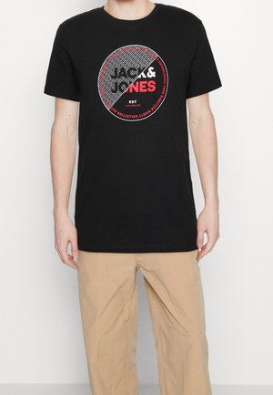 Person trägt ein schwarzes T-Shirt mit einem großen runden Jack & Jones-Logo in Rot und Weiß, kombiniert mit beigen Hosen.
