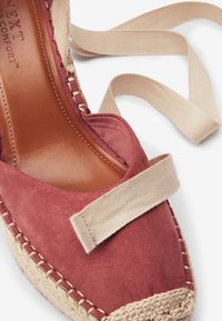 Espadrille en suède rouge avec une sangle en tissu beige et une semelle en jute tissée. Présente un intérieur en cuir lisse et des détails cousus le long du bord.
