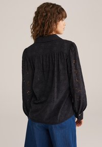 Črna čipkasta bluzica s ovratnikom, napihnjenimi rokavi in pregibnim hrbtom. Vsebuje cvetlične vzorce in poudarjene zapestnice s gumbi.