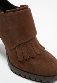 Stivaletto in suede marrone con un'accento frangiato nella parte anteriore, forma sagomata e suola in gomma per una migliore aderenza. Texture liscia.