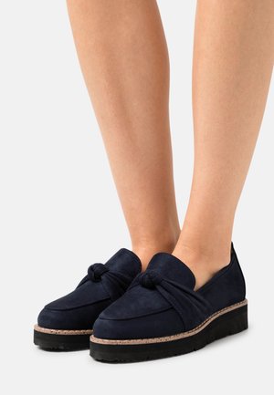 Mocassins en suede bleu marine avec un détail en nœud à l'avant, dotés d'une épaisse semelle noire et d'une semelle intermédiaire texturée pour un aspect robuste.