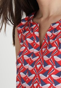 Rotes ärmelloses Oberteil mit einem Vogelprint in Weiß und Blau, mit V-Ausschnitt und Knopfdetails an der Vorderseite. Weicher Stoff, geometrisches Muster.