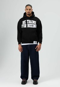 Zwarte hoodie met witte tekst "HE TRAINS FOR BOXING," voorzak, gecombineerd met losse blauwe spijkerbroek en zwarte schoenen. Simpel ontwerp, casual pasvorm.