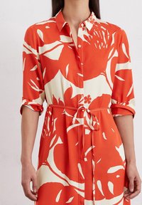 Donna che indossa un vestito a maniche corte con bottoni, a stampa floreale rosso e bianco, con le maniche arrotolate e una cintura annodata in vita.