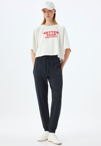 Witte kortgeknoopt t-shirt met rode tekst "BETTER THAN YESTERDAY," gecombineerd met zwarte tapered broek en witte sportschoenen. Eenvoudig ontwerp; casual pasvorm.