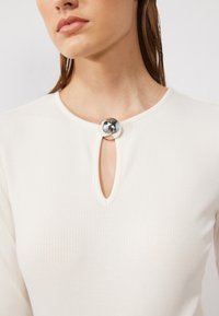 Ribbad långärmad topp i creme med en nyckelhålsutskärning, som har en framträdande metallknapp vid stängningen för extra detalj.