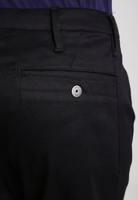 Černé kalhoty s hladkým povrchem, s zadní kapsou zabezpečenou kovovým knoflíkem. Jednoduchý design bez vzorů nebo akcentů.