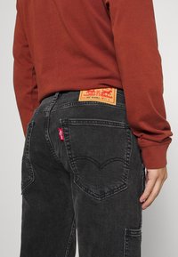 Jeans de denim negro con un diseño de bolsillo trasero en forma de corazón, etiqueta de logo roja y un parche de cuero marrón en la cintura.