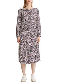 Femme portant une robe midi à manches longues bleu marine à fleurs, associée à des baskets blanches, debout devant un fond blanc.