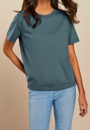 Vrouw draagt een effen teal T-shirt met korte mouwen en lichtblauwe spijkerbroek, met één hand op haar heup tegen een beige achtergrond.