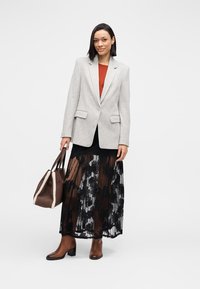 Grijze blazer over een roestrood topje, zwarte kanten rok met bloemmotief, bruine handtas en bruine laarzen met hak. Eenvoudige achtergrond.