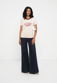 Ανοιχτό μπεζ βαμβακερό t-shirt με κόκκινο γραφικό "GUESS" και μπορντό περιθώριο, συνδυασμένο με ψηλόμεσα φαρδιά τζιν σε σκούρο χρώμα.