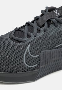 Svart mesh-sportskor med en texturerad design, gummisulor, platta snören och en diskret logotyp präglad på sidan.