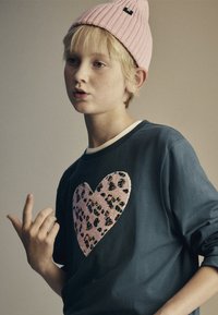 Kék pulóver nagy, rózsaszín leopárdmintás szívmintával, kerek nyakkivágással és hosszú ujjakkal. Egy világos rózsaszín kötött sapkával párosítva.