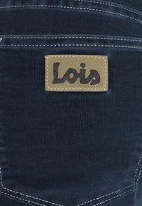 Tmavě modrá kordurová látka s béžovým koženým štítkem přišitém na kapse, na kterém je černým písmem vyšitý název značky "Lois".