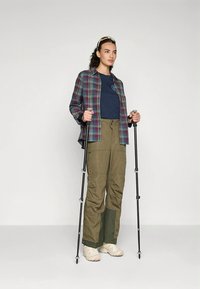 Koszula w kratę w odcieniach zieleni i niebieskiego, granatowy t-shirt, oliwkowe spodnie cargo, trzymająca kijki trekkingowe, nosząca jasne buty i okulary przeciwsłoneczne.