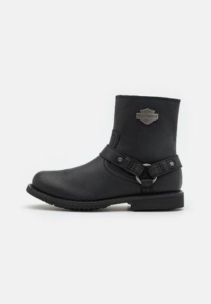 Botte de moto en cuir noir mi-mollet avec sangle de harnais, détail en anneau métallique et logo Harley-Davidson sur la tige.