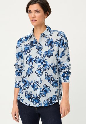 Kobieta w jasnoniebieskiej koszuli z długim rękawem w ciemnoniebieski kwiatowy wzór i ciemnych dżinsach, stojąca z rozluźnionymi ramionami na jednolitym tle.