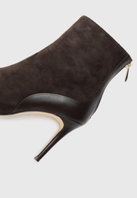Botín de ante marrón de tacón alto hasta el tobillo con un acento de cuero negro brillante y un detalle de cremallera dorada en la parte trasera.