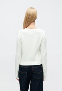 Fehér, texturált pulóver kerek nyakkal és hosszú ujjakkal, sötétkék farmerrel, amelyen varrott hátsó zsebek találhatók.