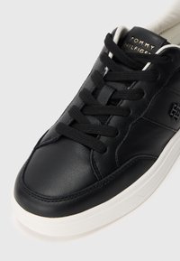 Černé kožené tenisky s bílou podrážkou, černými tkaničkami a zlatým nápisem "Tommy Hilfiger" na jazyku.