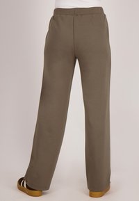 Persoon in losse, olijfgroene broek en bruine sandalen met dikke zolen, staand tegen een effen, lichte achtergrond.