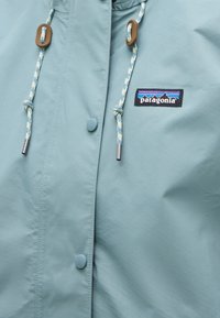 Μπροστινή όψη ανοιχτό μπλε μπουφάν Patagonia με κουμπιά στερέωσης, ρυθμιζόμενα λευκά και μπλε κορδόνια με καστανά κλείστρα και λογότυπο της μάρκας στο στήθος.