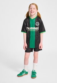 Svart och grön sporttröja med vertikala ränder och en logotyp, kombinerad med svarta shorts och gröna sneakers med vita detaljer och kardborreband.