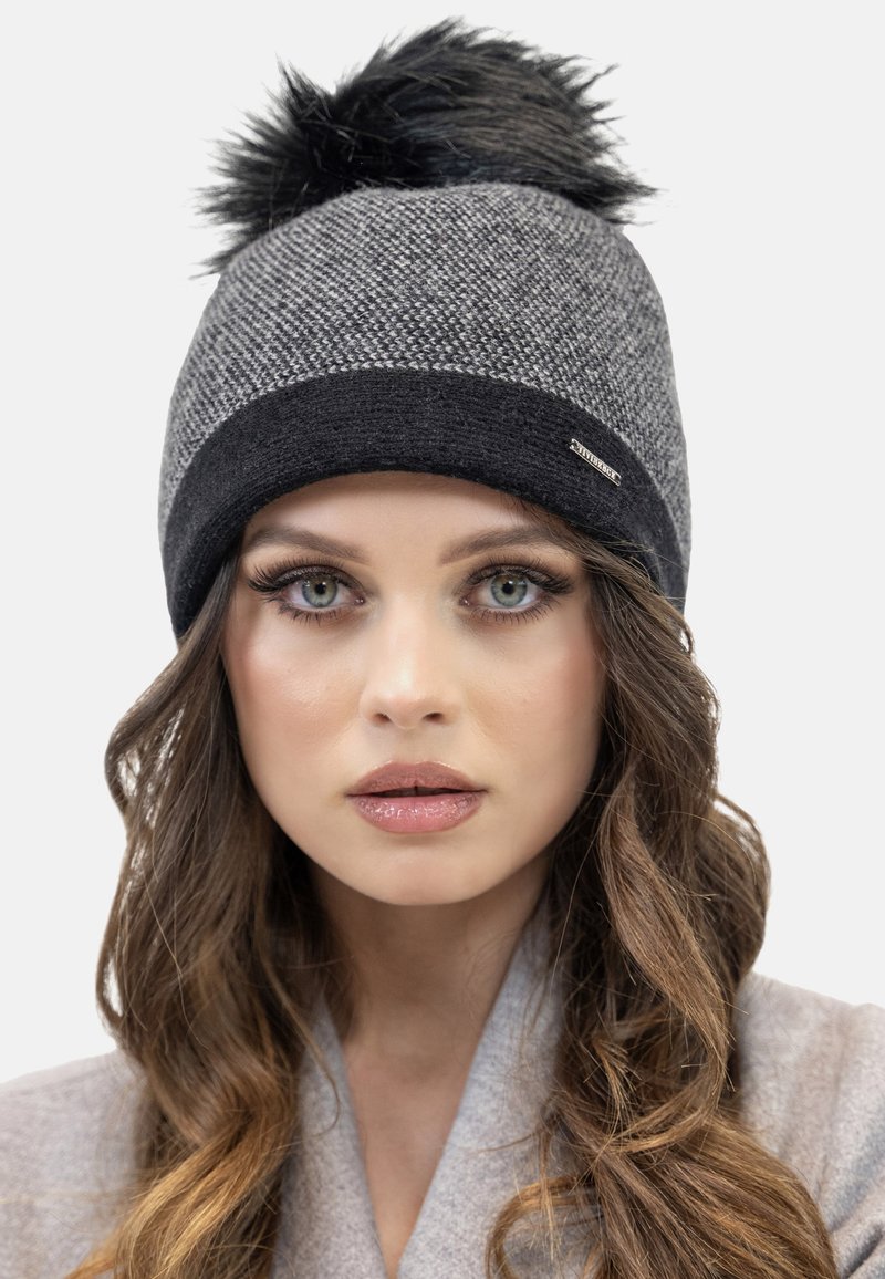 Gorro de punto en gris con un pom-pom de pelo sintético negro. Presenta un patrón texturizado y una banda negra en contraste en la base.