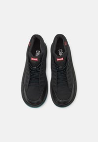 Black sneakers with a textured fabric upper, featuring blue accents, flat laces, and a smooth rubber sole. "Camper" logo on the tongue.