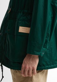 Λεπτομέρεια στο μανίκι και την τσέπη ενός πράσινου μπουφάν Barbour Paul Smith με το χέρι ενός ατόμου να ακουμπάει δίπλα σε μπεζ παντελόνι.